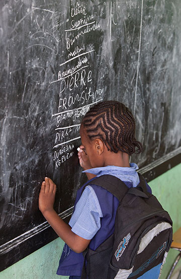 A child writing on a chalkboard with a backpack on.