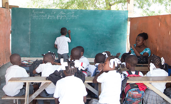 A picture of the children in an outdoor classroom, with a child writing on the chalkboard.